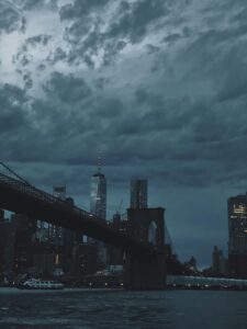 A dramatic view of the Brooklyn Bridge and New York City skyline under dark, moody clouds at dusk.